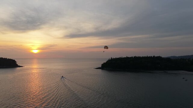 A parasail floats above the ocean as a boat moves steadily across the water, with a beautiful sunset illuminating the sky and reflecting on the calm sea near a lush green island