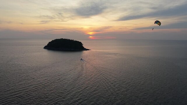 A serene sunset view over the ocean features a small island and a parasailer gliding above the water, while a boat moves steadily towards the horizon, creating a tranquil coastal scene