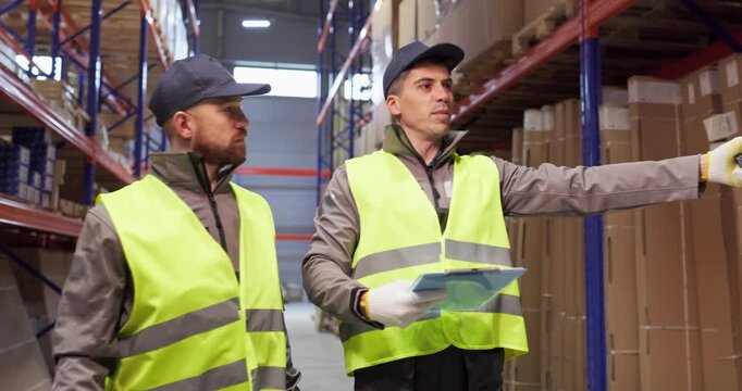 Workers in warehouse conduct inventory inspection with radio. Two staff in vests check shelves and boxes, handling logistics with teamwork and gloves. Clear reliable warehouse teamwork concept.