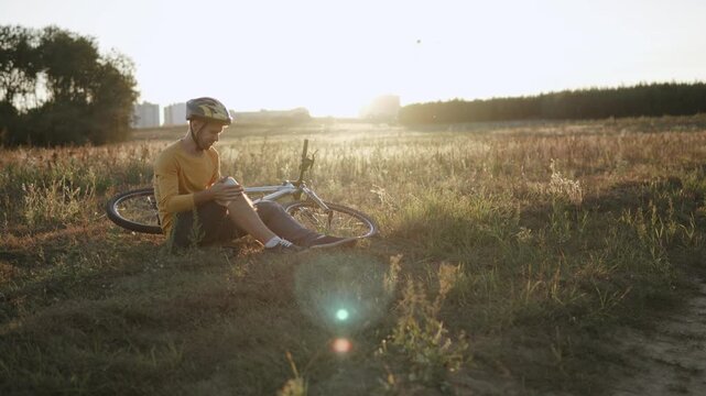 Caucasian man sits with bandaged knee in field near bicycle and winces in pain. Accident and self help during bike ride. Person with insurance claim due to injury from active lifestyle.