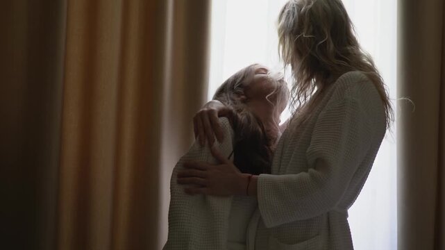 A happy family weekend spent together. Early in the morning, a woman hugs and kisses her daughter while standing by the window in white waffle robes. High quality FullHD footage
