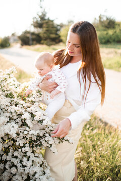 Happy young woman 30-35 year old holding baby over flowers in sunny day outdoors. Summer vacation season. Motherhood.