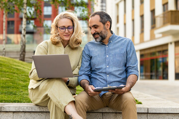 Fototapeta premium Colleagues discussing work on laptop while sitting outdoors in urban plaza