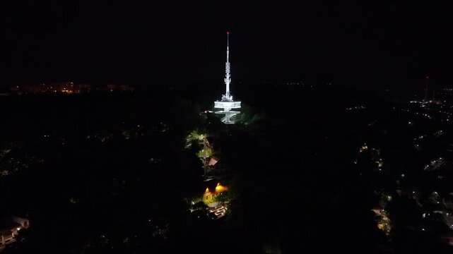 Night drone footage over Miskolc, Hungary, featuring the iconic Avas TV Tower rising above the city skyline. The video captures the illuminated tower, glowing streets, hillside neighborhoods
