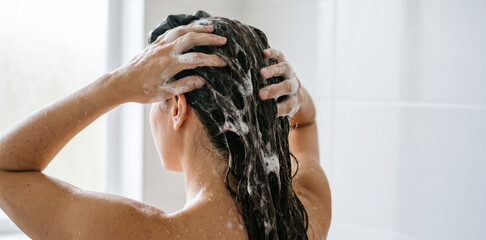 Naklejka premium A woman stands in a shower, applying shampoo to her hair. Water runs over her body as she massages her scalp. The bathroom is bright with natural light coming through the window