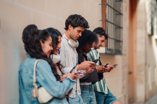 Diverse group of young friends focused on their smartphones, ignoring each other while standing against a wall