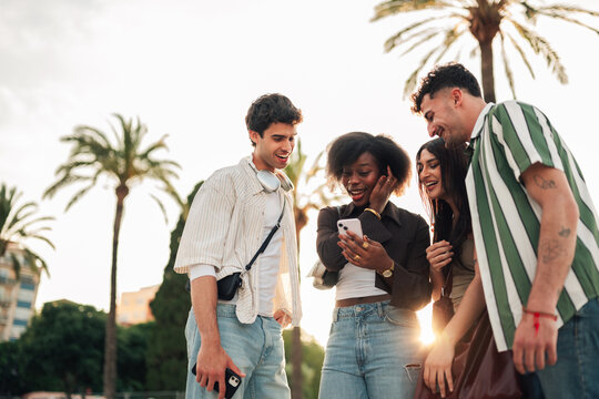 Diverse young friends smiling and looking at a smartphone screen, sharing a moment and enjoying a digital connection outdoors