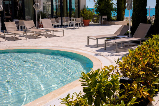 Outdoor hotel pool with curved blue water edge, dark gray sun loungers and closed umbrellas on tiled deck, large white resort building on a sunny summer day