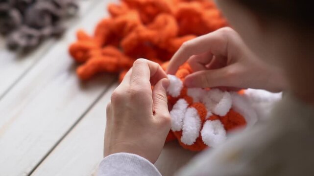 Little baby's fingers are knitted using white and orange plush yarn.. Children's hobby. The girl sits holding a woolen thread in her hands.