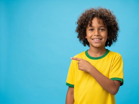 Happy young boy with curly hair wearing a yellow and green t-shirt pointing his finger to the side against a bright blue background, providing ample copy space for advertising and marketing
