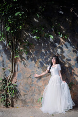 Fototapeta premium Young Asian Woman in White Dress Standing by Rustic Stone Wall with Natural Light