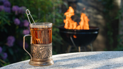 A vintage metal tea glass holder with ornate grapevine engravings stands on a garden table. In the background, a barbecue grill glows with orange flame beside blooming rhododendron bushes © Anya Kamyshnikova