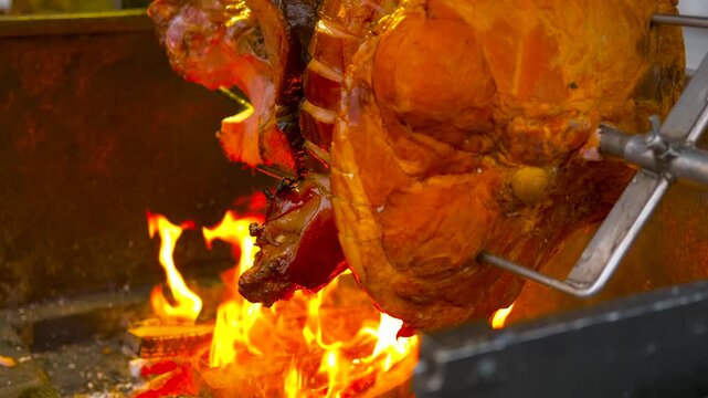 Juicy pork leg roasting on a spit over open fire. Large juicy pork knuckle roasting on a rotating spit over a wood fire at a street food market. Delicious traditional meal cooking outdoors