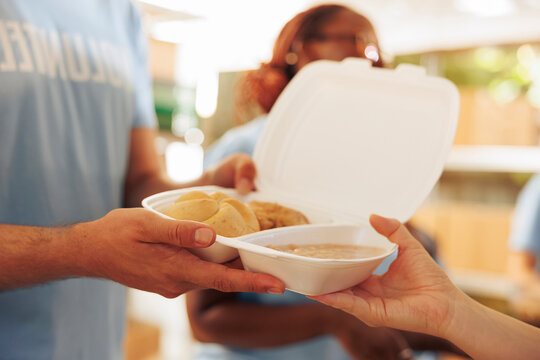 Less fortunate woman receiving free food from male volunteer at outdoor food bank; a pair of caucasian hands holding meal box donation for the hungry and homeless people.