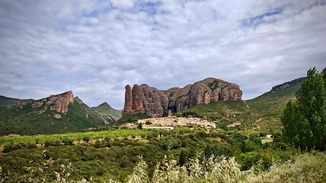 Aguero, Spain, is shown nestled at the base of the Mallos de Aguero rock formations. The village buildings are surrounded by lush greenery, blending into the verdant landscape. 
