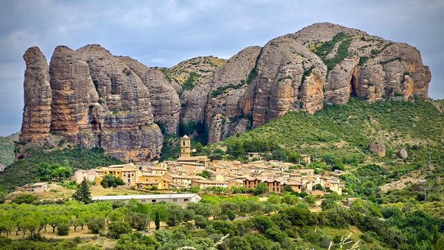 A picturesque view of the village of Ag&uuml;ero in Spain, positioned at the base of the impressive Mallos de Ag&uuml;ero rock formations. The rocks are large, vertical cliffs with a rugged texture 