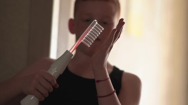 A teenager uses a Darsonval device. The young man runs his hand over the glass attachment designed for hair. Professional skincare at home. Teenage skin issues during hormonal changes