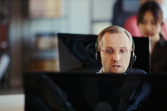 Middle aged Caucasian man wearing headset, assisting customers and monitoring computer in call center office. Ideal for customer support, help desk, outsourcing, service team marketing
