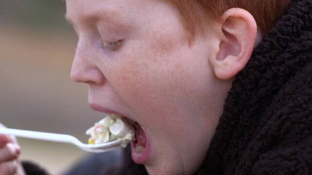A red-haired teenager enjoys a piece of salad from a disposable utensil in a park on a cloudy day. A casual al fresco lunch and a moment in everyday life. High quality FullHD footage