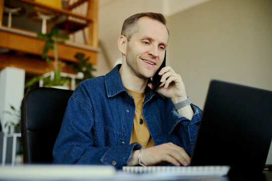 Young adult Caucasian man speaking on smartphone and typing on laptop in office, handling customer support and communication. Ideal for call center, remote work, business service