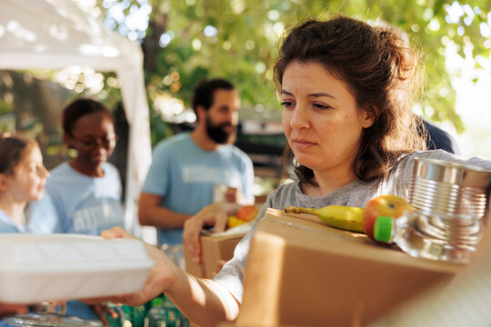 Volunteers hand out packages and meal boxes offering free food to the homeless and needy; underprivileged caucasian woman receiving hunger relief donation package from charity workers.