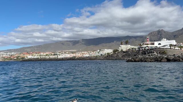Scenic view of the Tenerife coastline from a moving motorboat on the Atlantic Ocean. Resort buildings, rocky shoreline and volcanic mountains under clouds, coastal tourism, boating adventure, ocean tr
