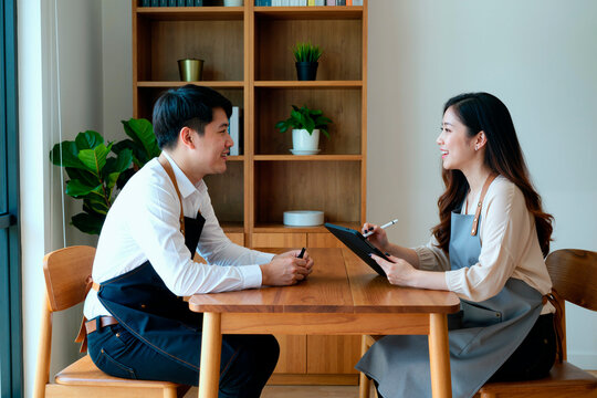 Asian young adult cafe owners discussing business plan at table, reviewing notes on tablet during meeting. Useful for small business, partnership, startup marketing concepts