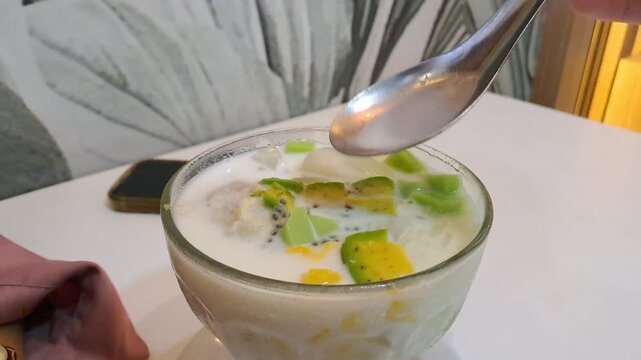 Close-up of an Asian woman enjoying a viral creamy Durian Es Teler in a cafe. The footage captures her stirring the refreshing Indonesian iced dessert filled with avocado, coconut, jackfruit, and prem