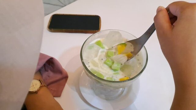Close-up of an Asian woman enjoying a viral creamy Durian Es Teler in a cafe. The footage captures her stirring the refreshing Indonesian iced dessert filled with avocado, coconut, jackfruit, and prem