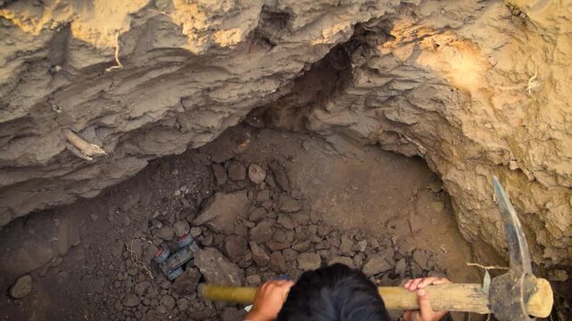 Young boy digging a deep pit with a pickaxe in rocky soil, manual excavation work and hard labor