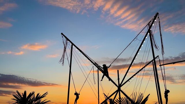 Slow motion: children silhouettes are jumping on bungee trampoline against warm dramatic sky at sunset at amusement park. Childhood, summer, fun and holiday concept