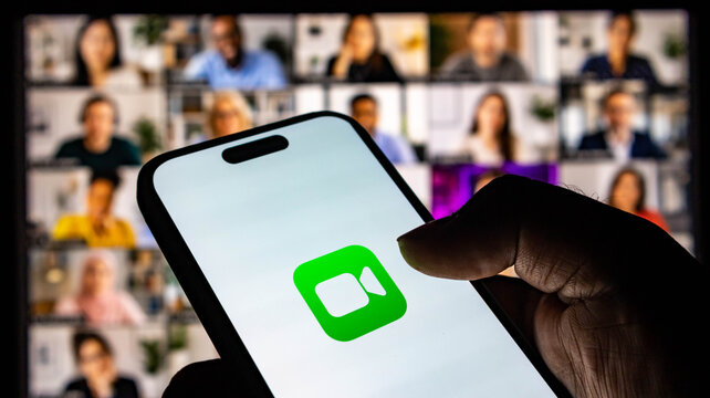 Person holding a smartphone displaying the facetime video call icon with a blurred group video conference in the background