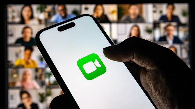 Closeup of a hand holding a smartphone displaying the facetime video call icon with a blurred group video conference in the background