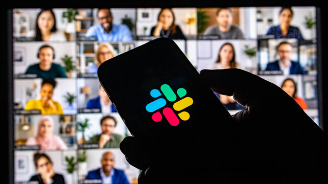 Person holding a smartphone displaying the slack logo in front of a computer screen showing a video conference call