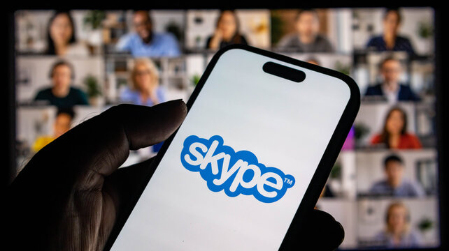 Closeup of a hand holding a smartphone displaying the skype logo with a blurred video conference call in the background