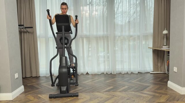 Woman exercises in reverse direction on elliptical trainer, using handles while facing forward. Home fitness routine for legs, coordination and varied indoor cardio.
