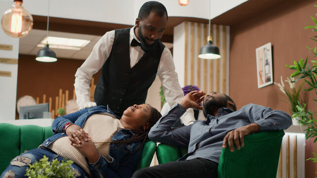 Hotel employee waking up people napping in lobby, inviting guests to check-in at hotel reception, exhausted couple travellers sleeping in lounge area while waiting for room.