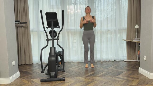 Woman in sportswear performs light vertical jumps on straight legs in living room near elliptical trainer and large window. Home fitness routine for lymph flow, rebound movement, morning activation