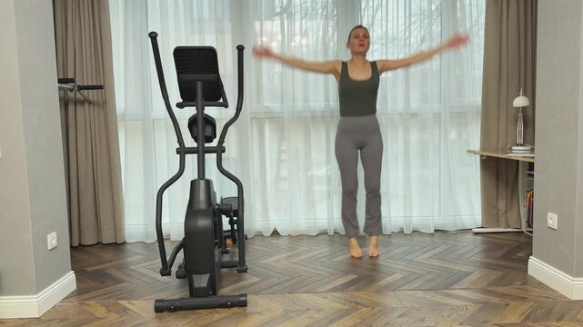 Female athlete performs jumping jacks with wide leg stance and overhead arm claps in living room. Classic home workout for full body activation and cardio training.