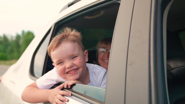 Smiling little boy sits in car with mother looks out window. Child son, smiles dreams, looking out open car window. Family trip with child. Happy family, kid enjoys drive in outdoors. Kid, mom plays