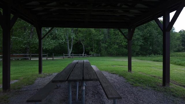 Drone flying through a covered picnic area in a freshly mowed field surrounded by a thick forest on a windy spring day