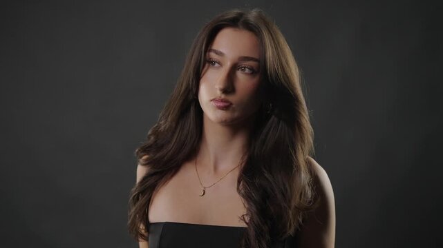 Hair care and beauty concept with a young woman posing and gently touching her long brown hair in a studio setting. This footage emphasizes glamour, fashion, and cosmetic themes, advertising hair prod