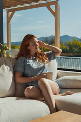 Obraz premium Redhead woman sitting comfortably on an outdoor sofa, holding a coffee mug and embracing a calm morning, enjoying the warm sunlight on her face with a mountain view