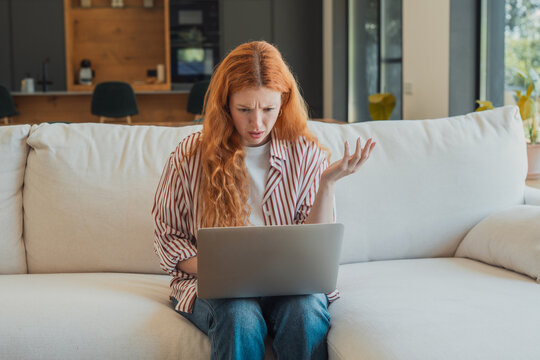 Frustrated redhead woman sitting on a couch at home, struggling with a laptop, finding difficulty and stress while dealing with technology confusion and a system glitched or unresponsive software