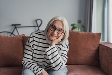 Obraz premium Senior woman with gray hair and glasses relaxing on a brown couch in her living room, smiling gently at the camera, peaceful and content in a casual, domestic setting