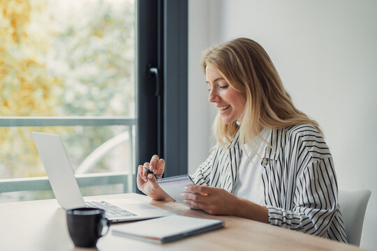 Young woman working productively from her home office, cheerfully looking at a laptop and holding a pen while taking notes in a spiral notebook by a window with natural light