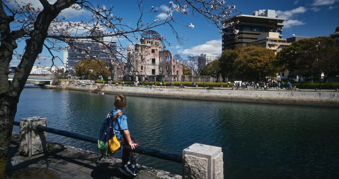 Child Sits On Rail And Looks Across River Toward Hiroshima Peace Memorial Dome - Wide, Slow Motion Shot