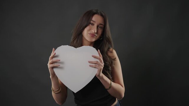 Love and affection concept with a young woman holding a white heart shape on a dark studio background. This shot romance and care, Valentine s Day themes and relationship content. The model s gentle e