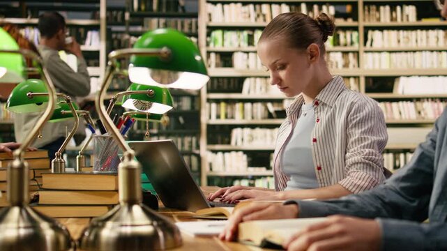 Young woman student working on laptop at library wooden table, peers study around green lamps and stacked books.