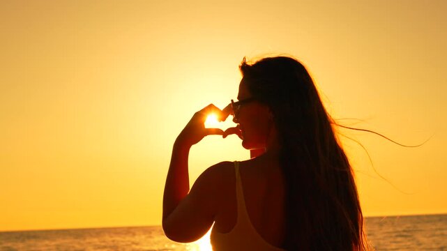 Silhouette heart sunset young woman makes a heart shape with her hands around the sun over ocean waves at the beach